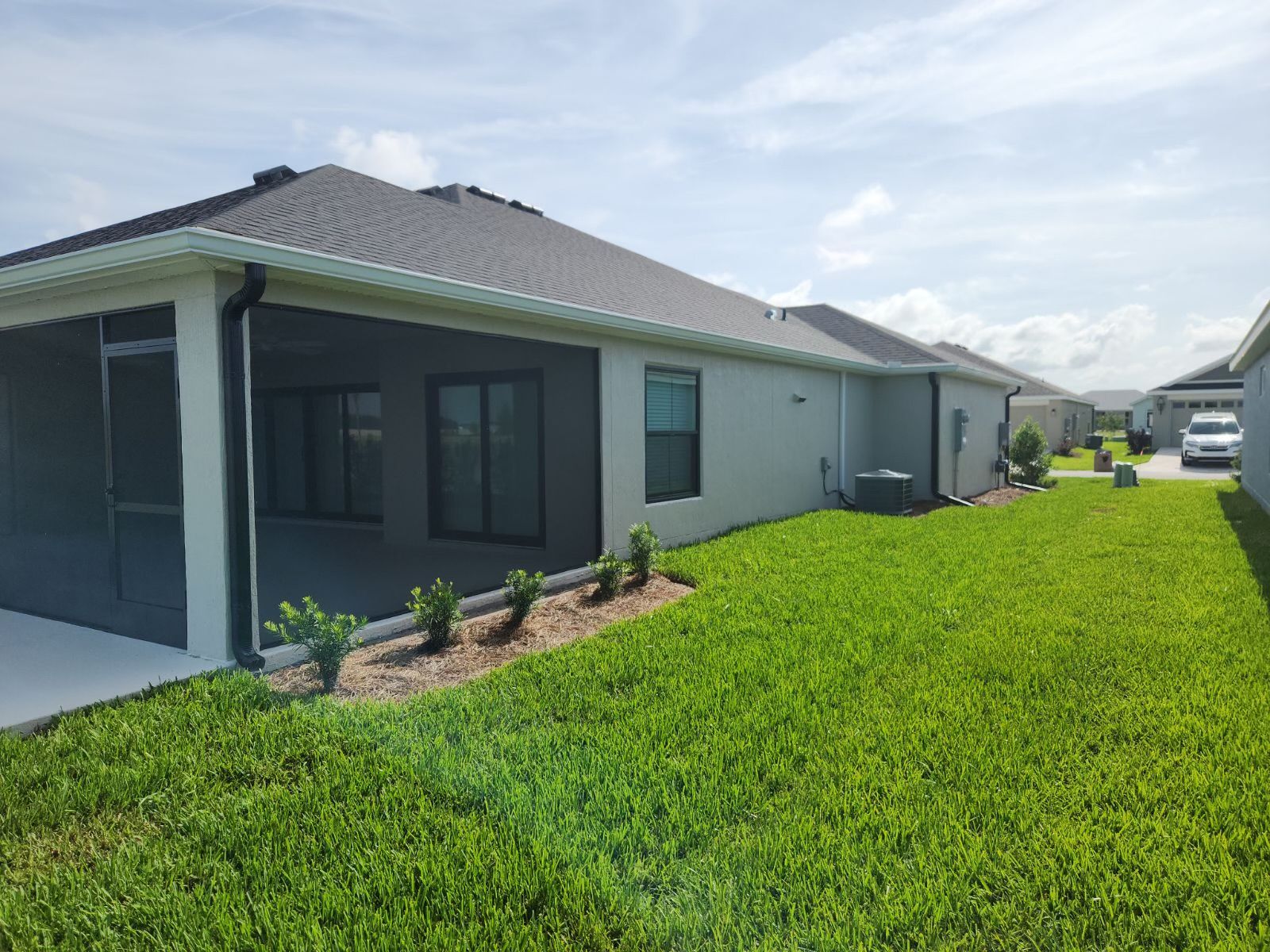 Backyard of a light-colored house with a screened porch, green lawn, and small bushes, under a blue sky.