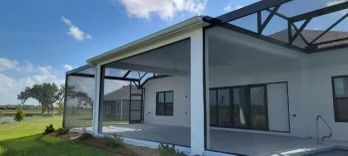 A screened-in patio of a white house with black accents on a sunny day; overlooking a green lawn and body of water.