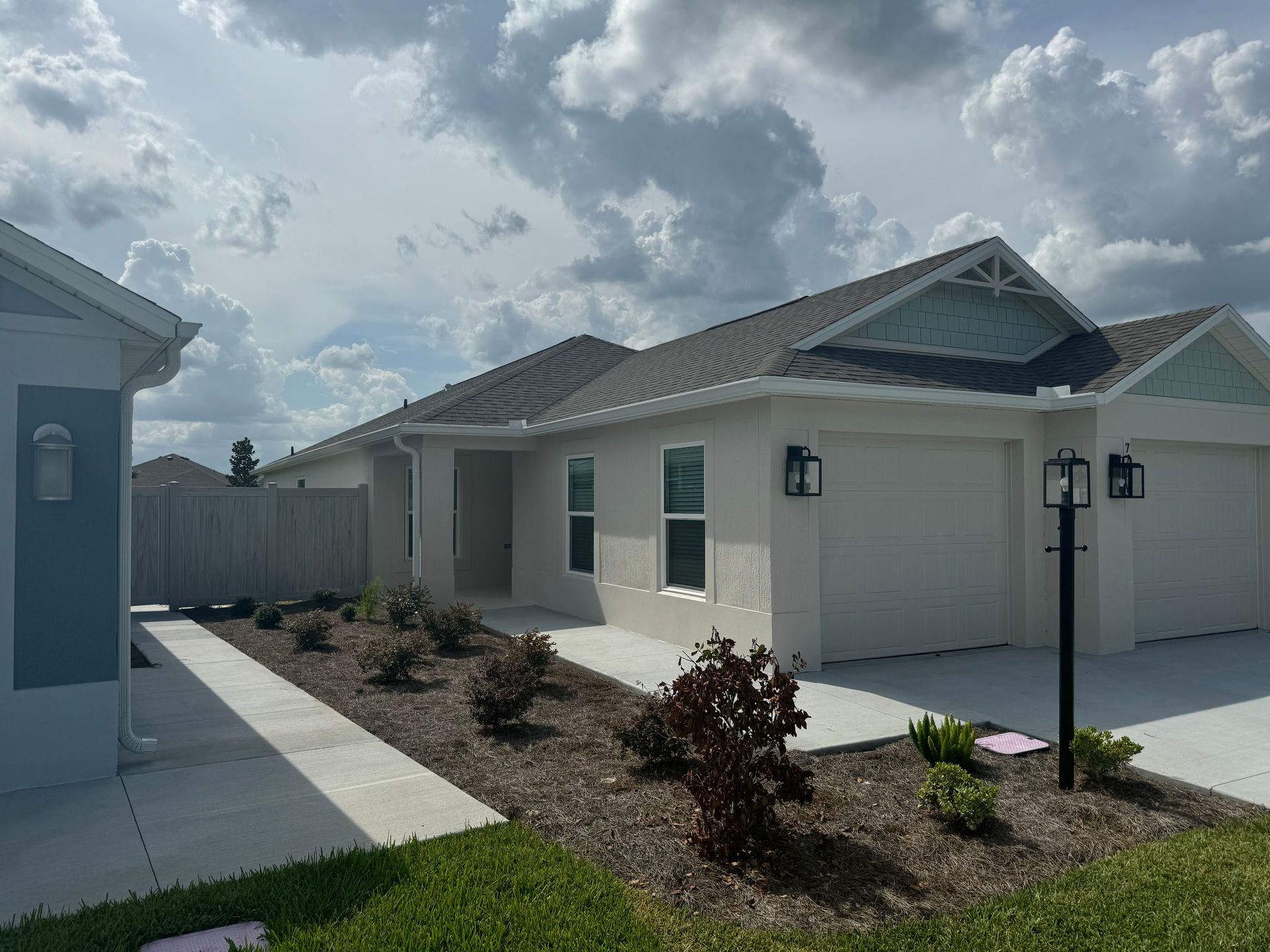 Light gray house with garage, black lamps, garden with plants, and cloudy sky.