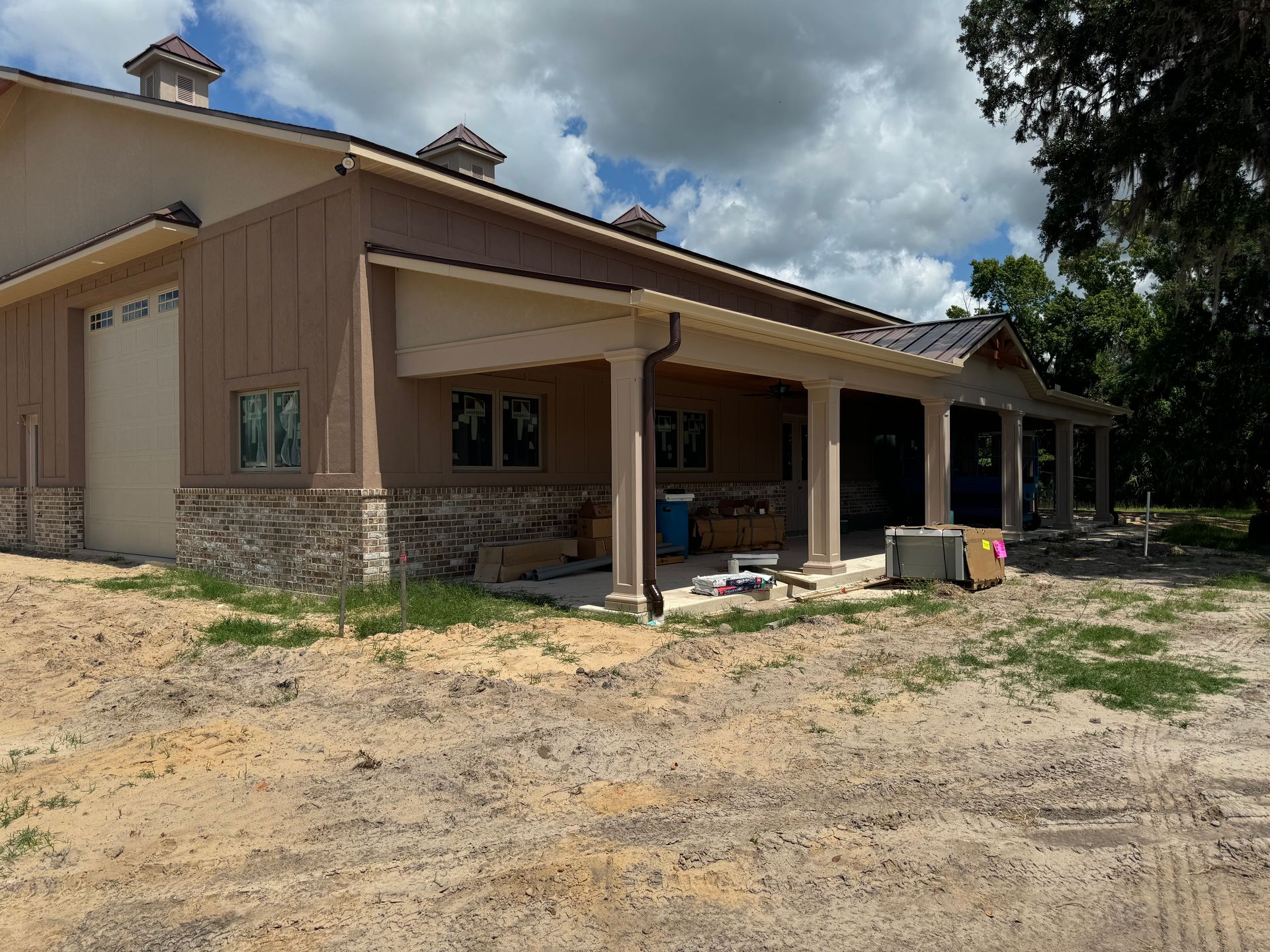 Building exterior under construction with a porch, beige stucco, brown accents, and a sandy yard.