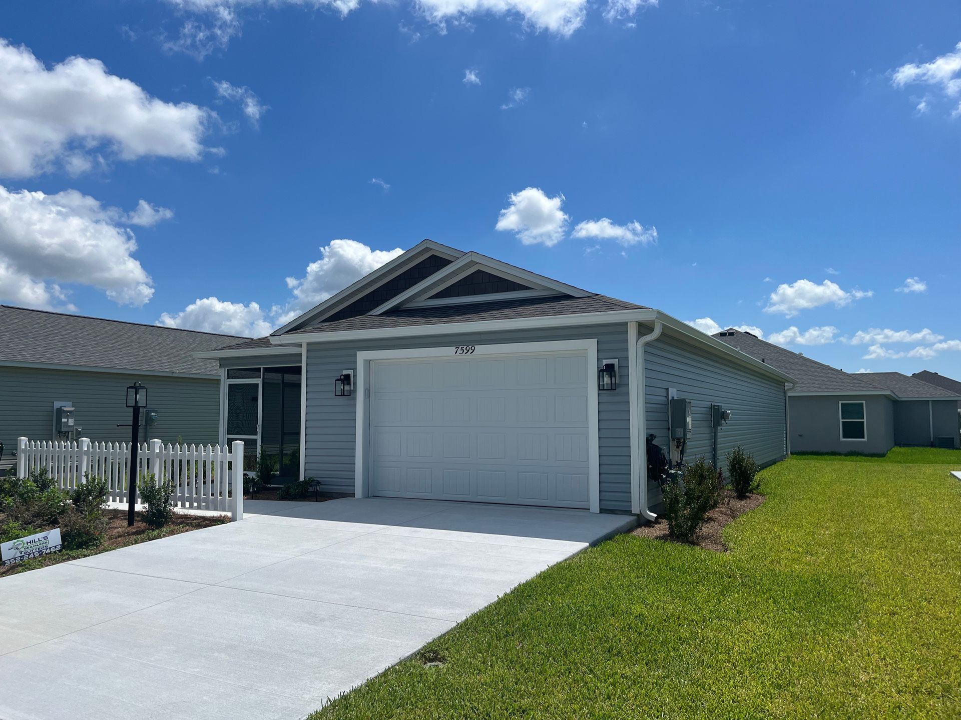 Blue-gray house with white garage door, driveway, and fence on a sunny day. Green grass and blue sky.