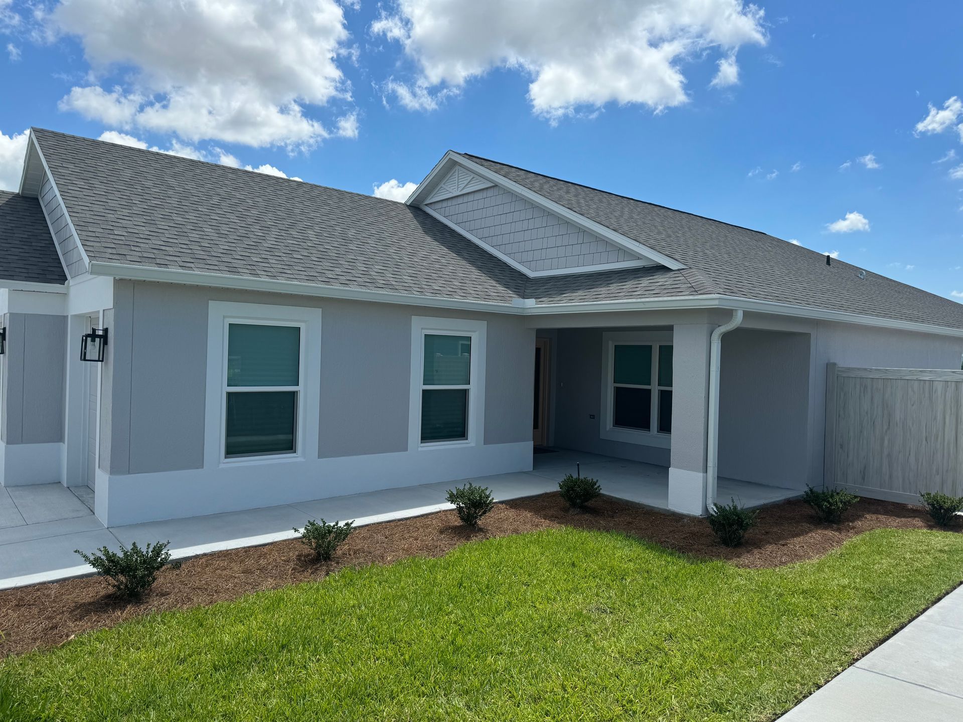 Gray-roofed, light gray house with a porch and green lawn under a blue sky with white clouds.