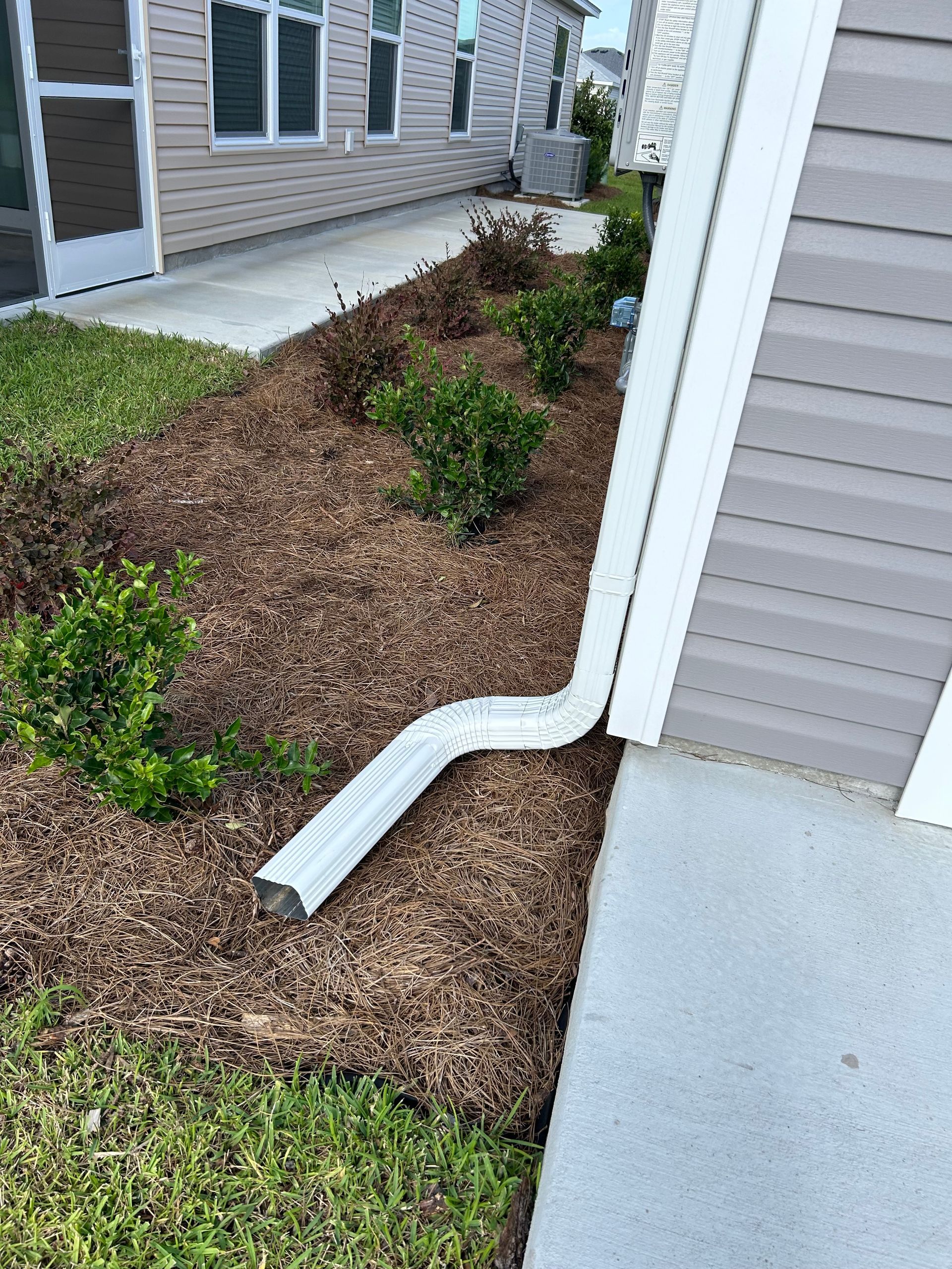 White gutter extending from a building to the ground, surrounded by mulch and greenery.