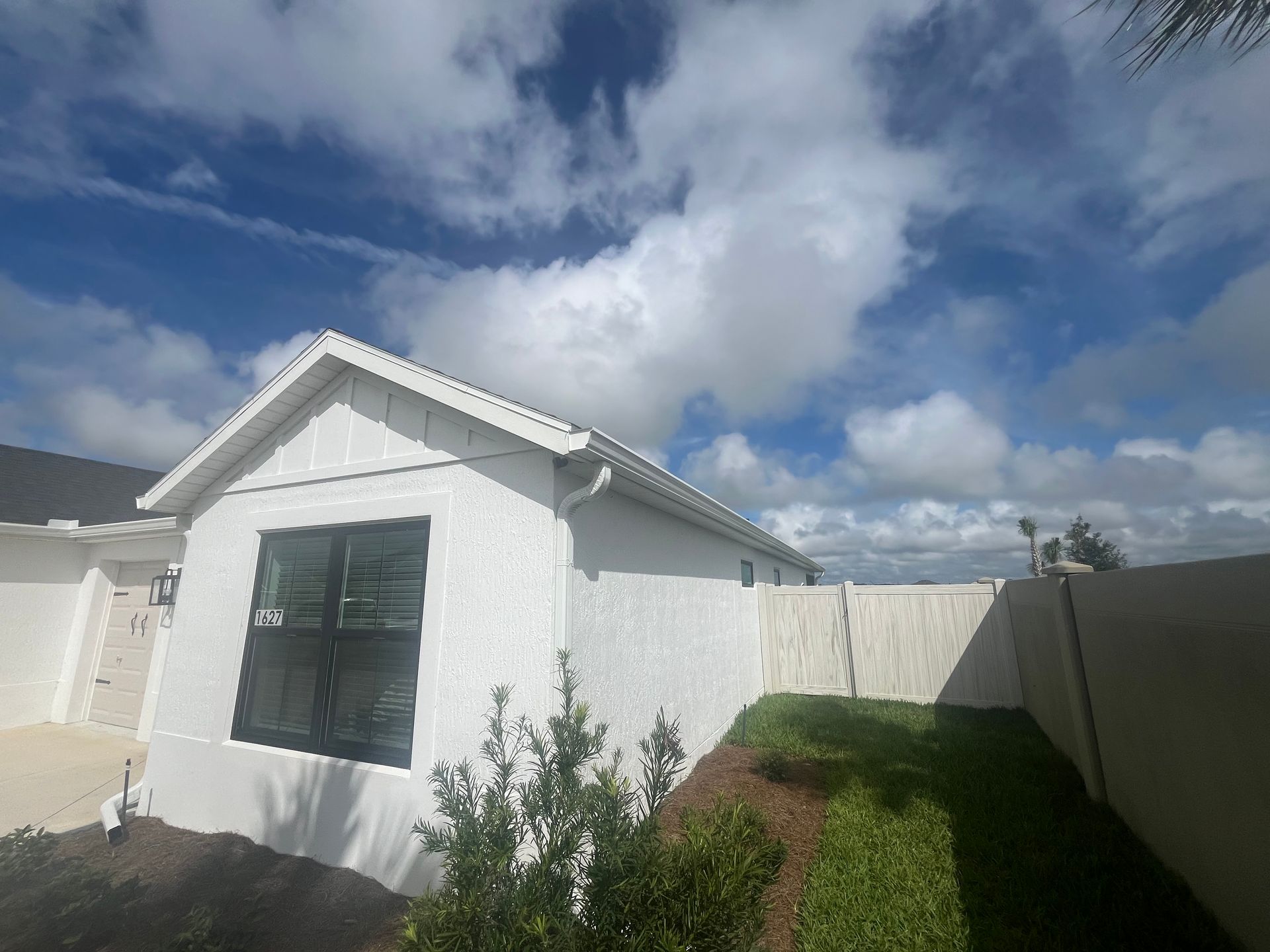 White house with a large window, surrounded by a fence and a blue sky with clouds.