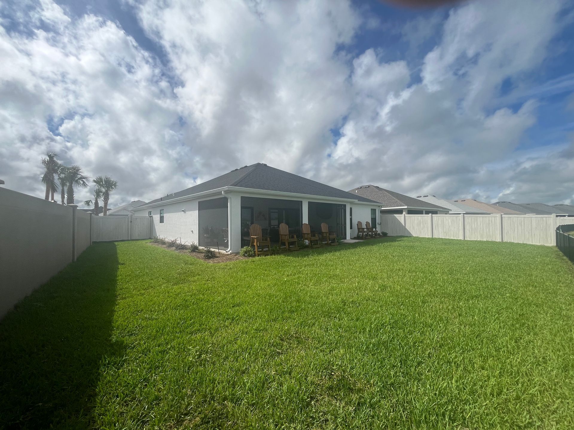 Backyard with green lawn, white fence, and house with screened patio under a cloudy sky.