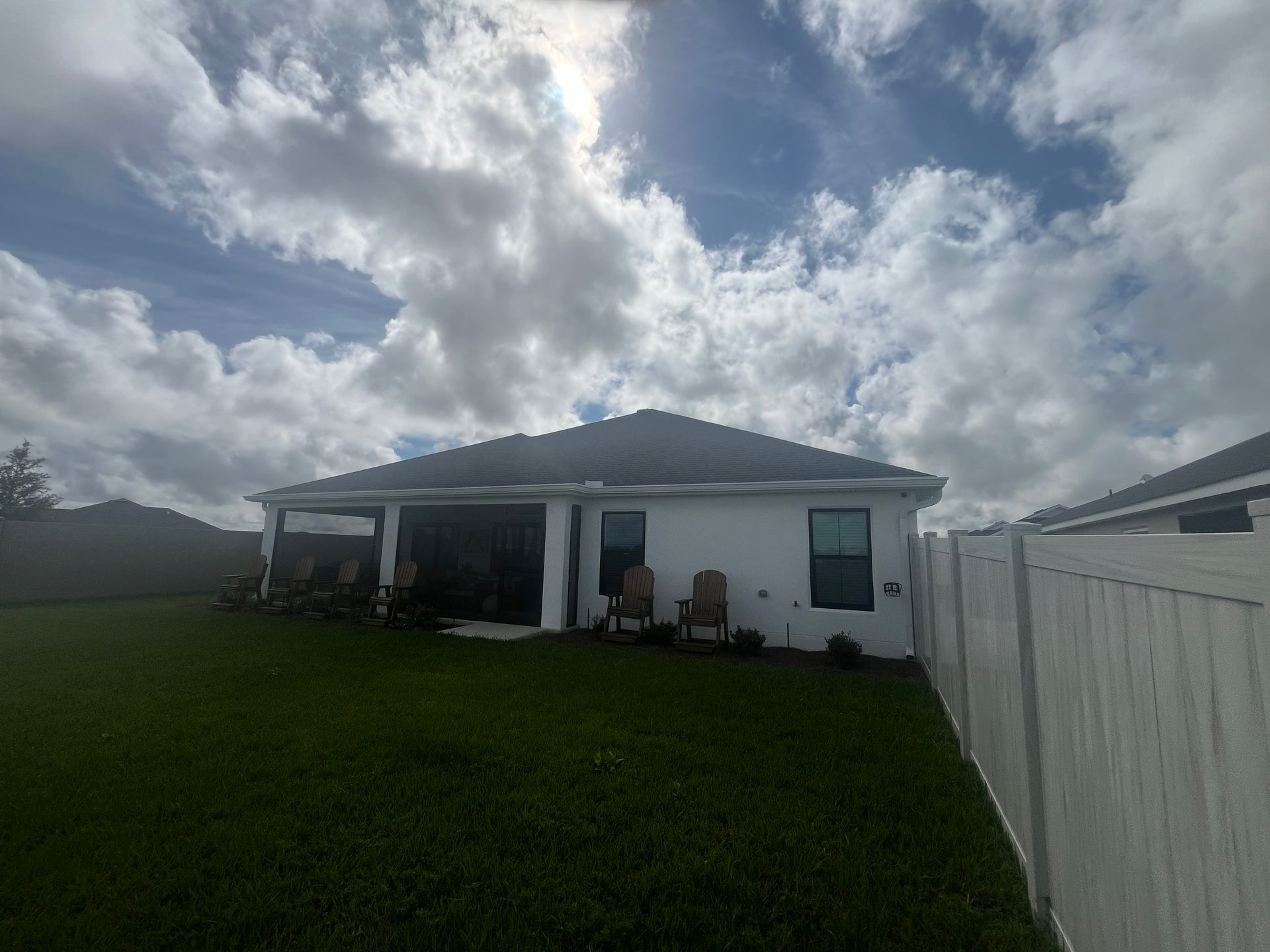 White house with dark roof, chairs on patio, green lawn, cloudy sky.