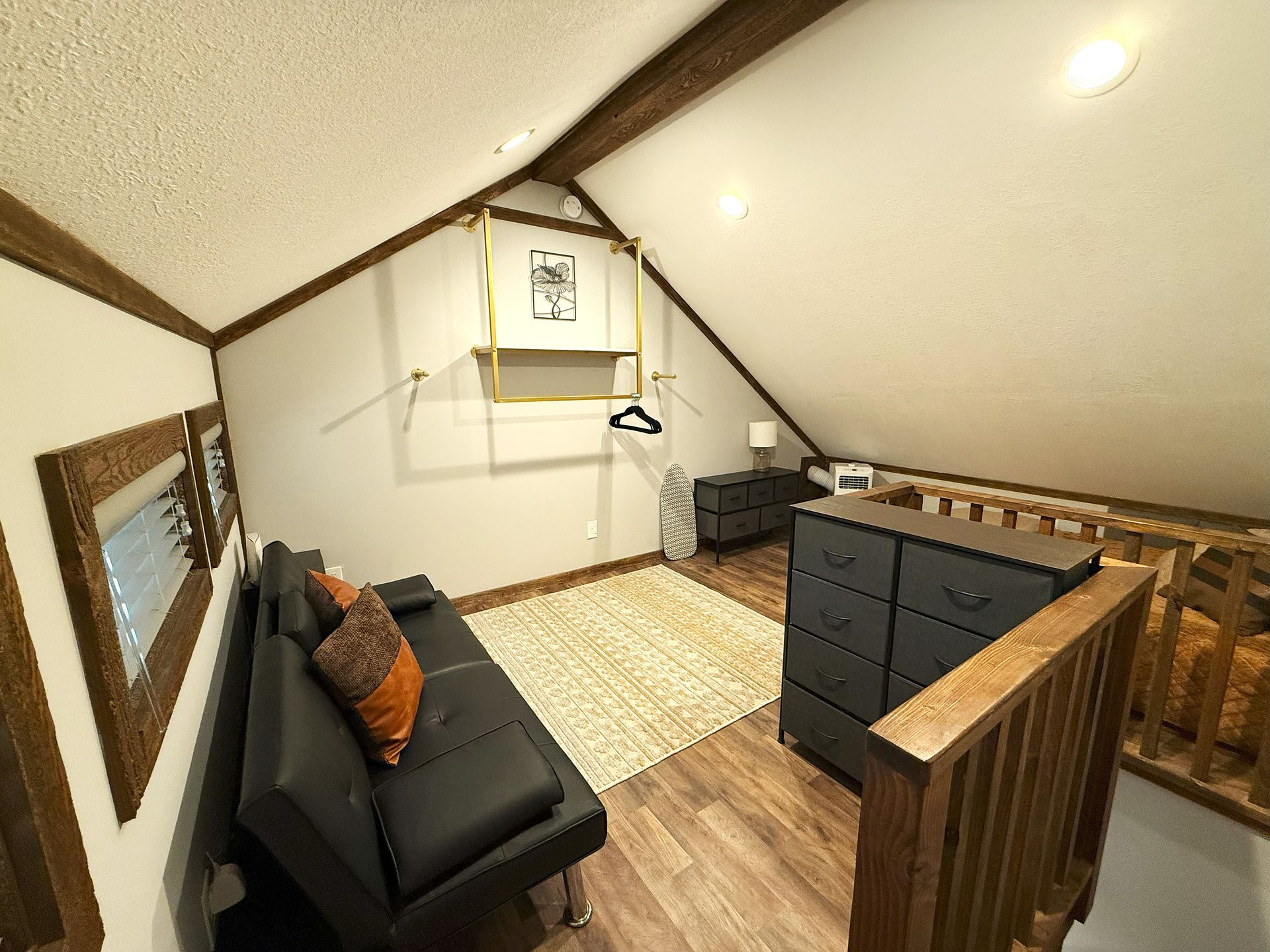 Loft bedroom with black futon, dresser, and light brown rug. Sloped ceiling with wood beams.