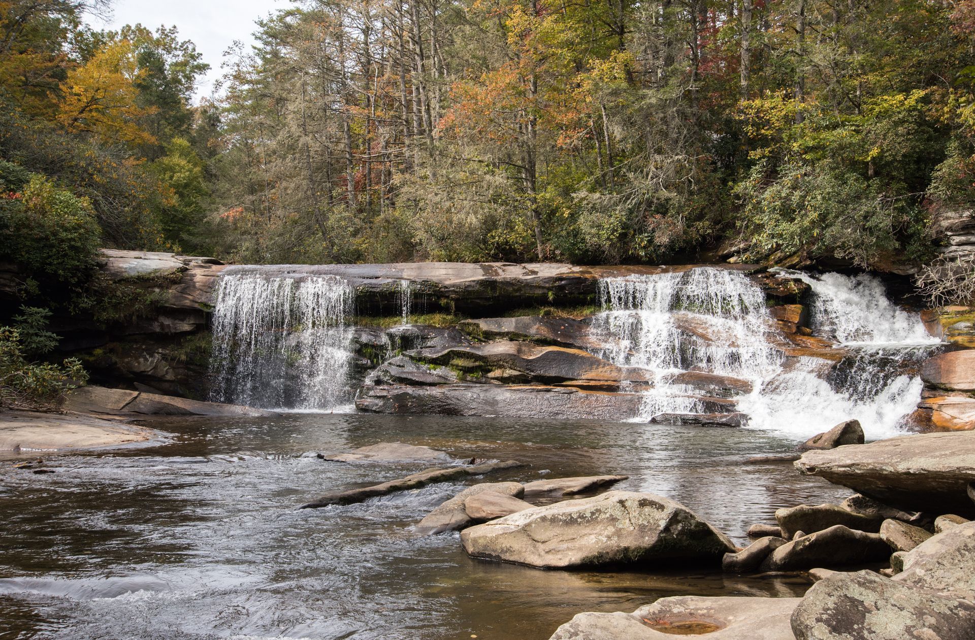 Waterfall cascading over rocks into a stream, surrounded by trees with fall foliage.