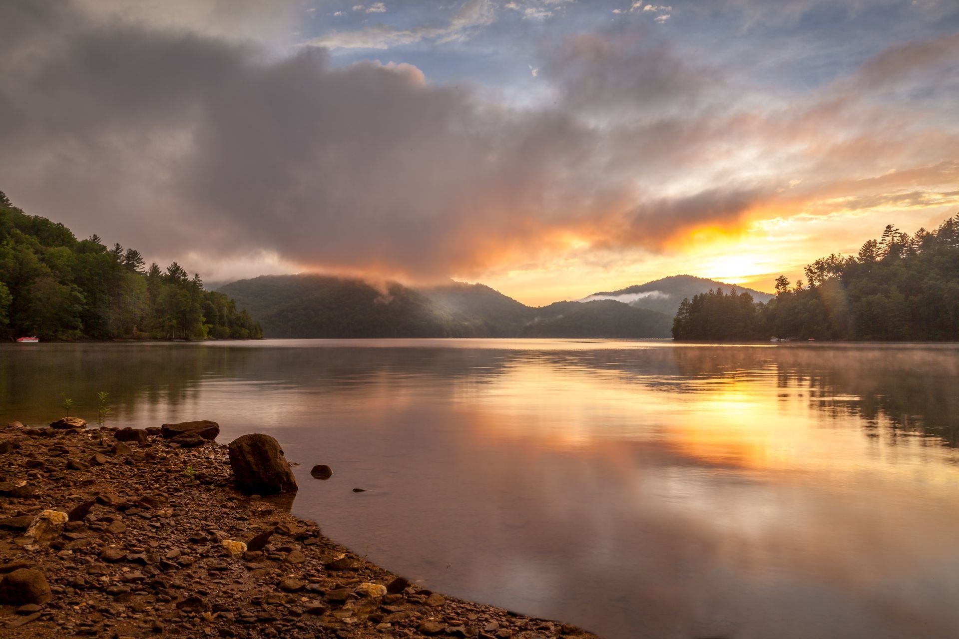 Sunrise over a calm lake reflecting golden light and cloudy sky, with forested hills on either side.