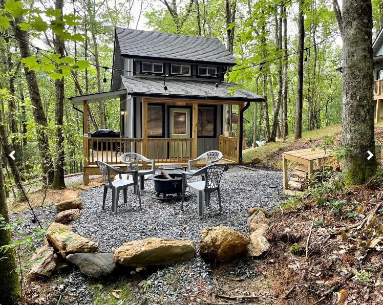 Small cabin in a wooded area with a porch, patio with fire pit, and outdoor seating.