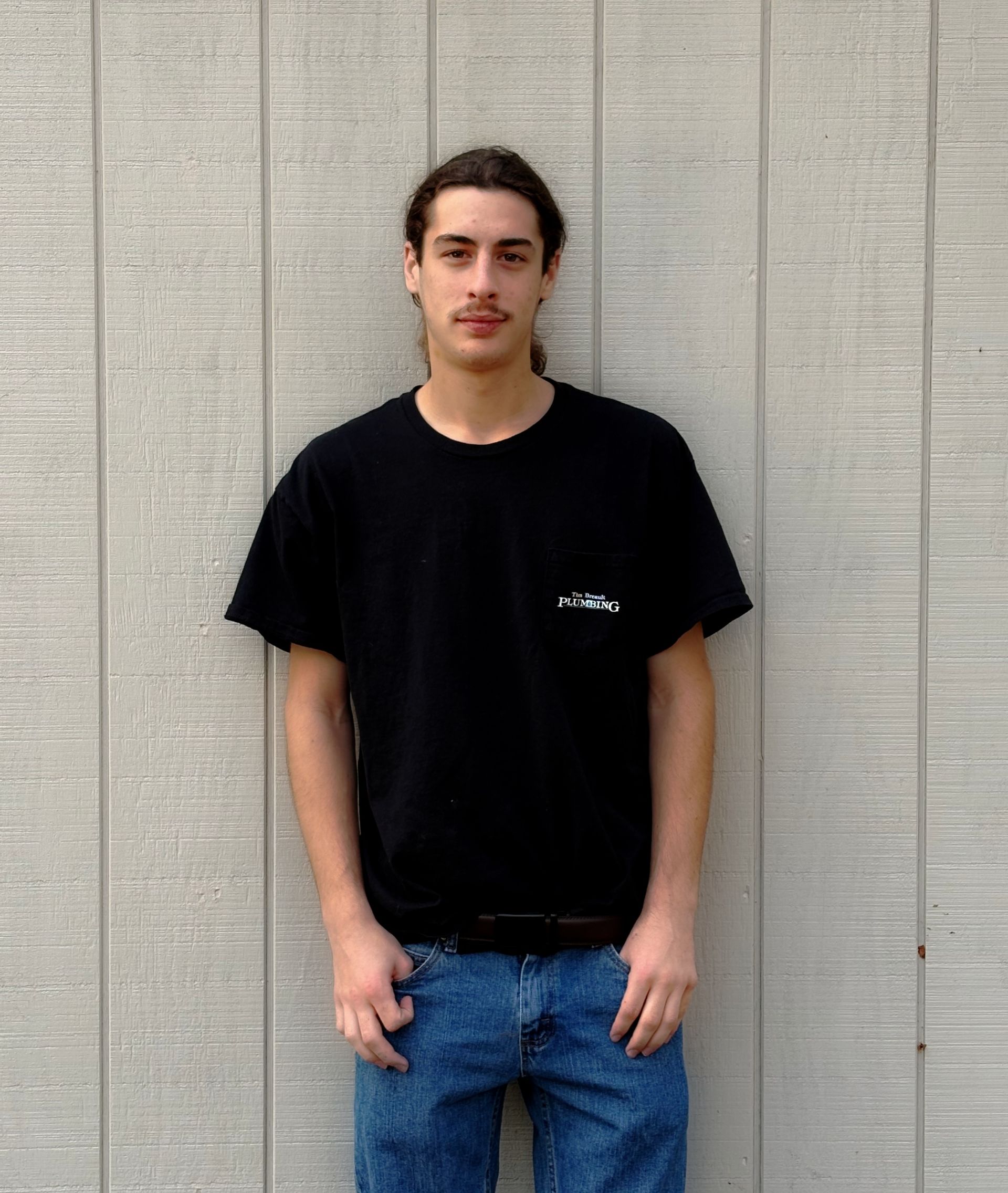 Bald man with crossed arms, wearing a grey t-shirt, smiling, standing outside in front of grey panels.