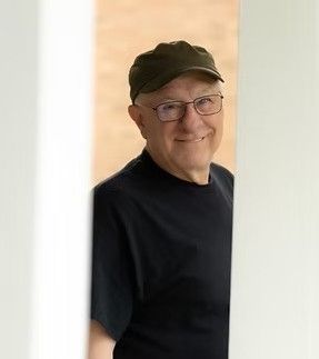 Man with glasses and green cap, smiling, framed by a white doorway.