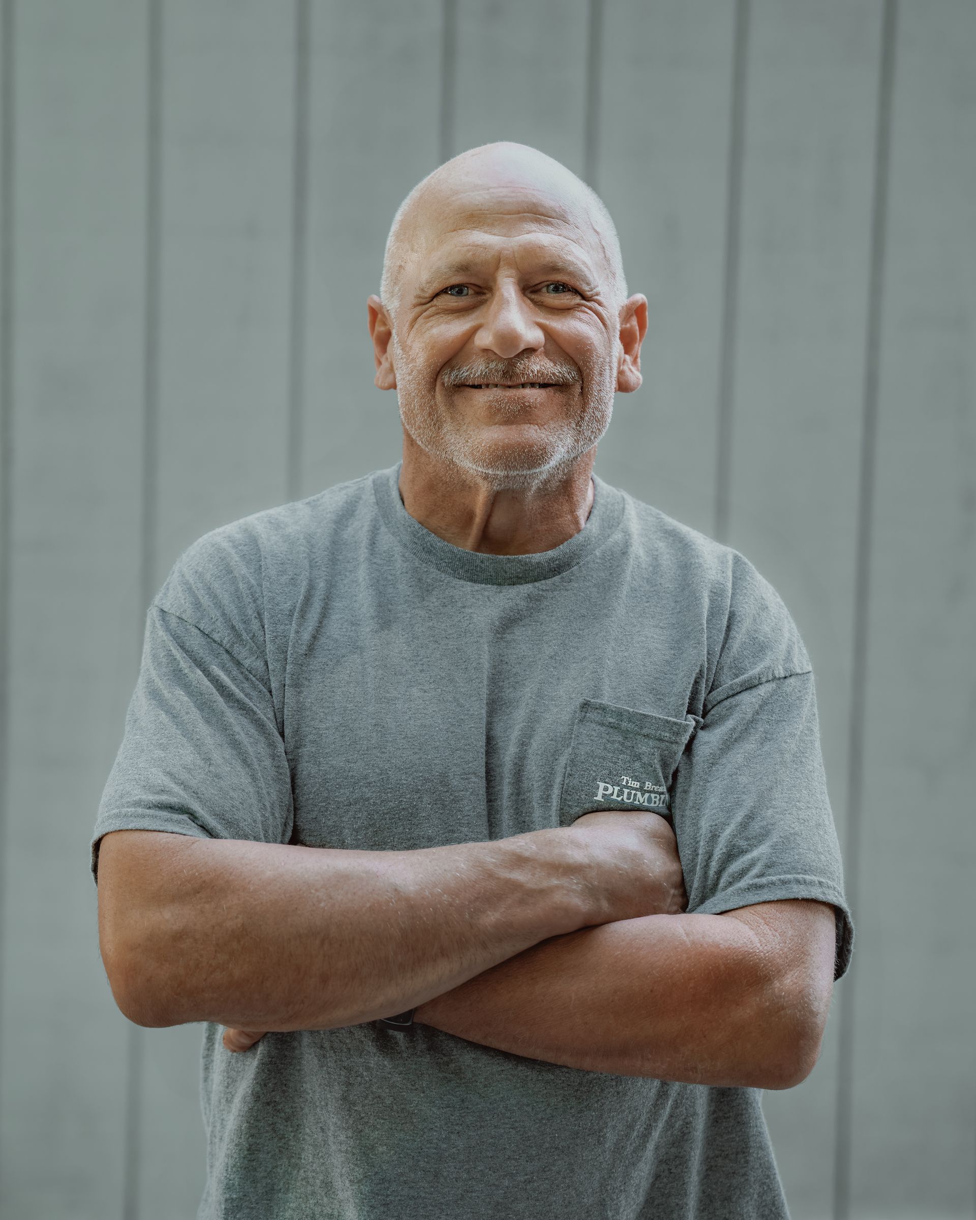 Bald man with crossed arms, wearing a grey t-shirt, smiling, standing outside in front of grey panels.