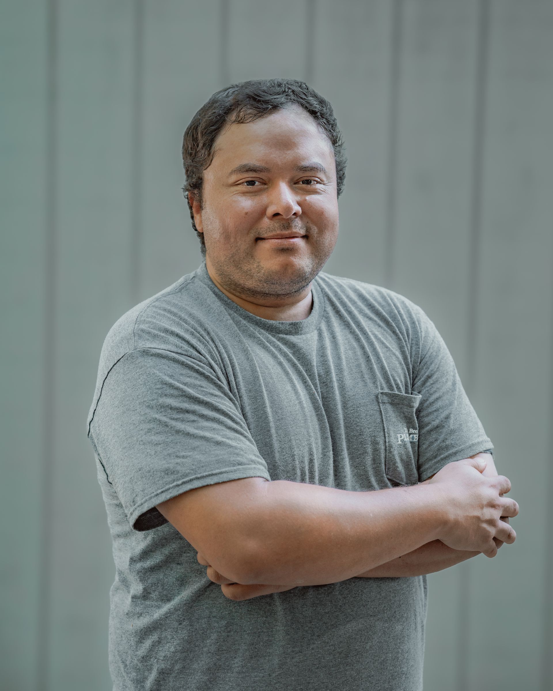 Man in gray t-shirt with arms crossed, standing in front of a light blue wall.