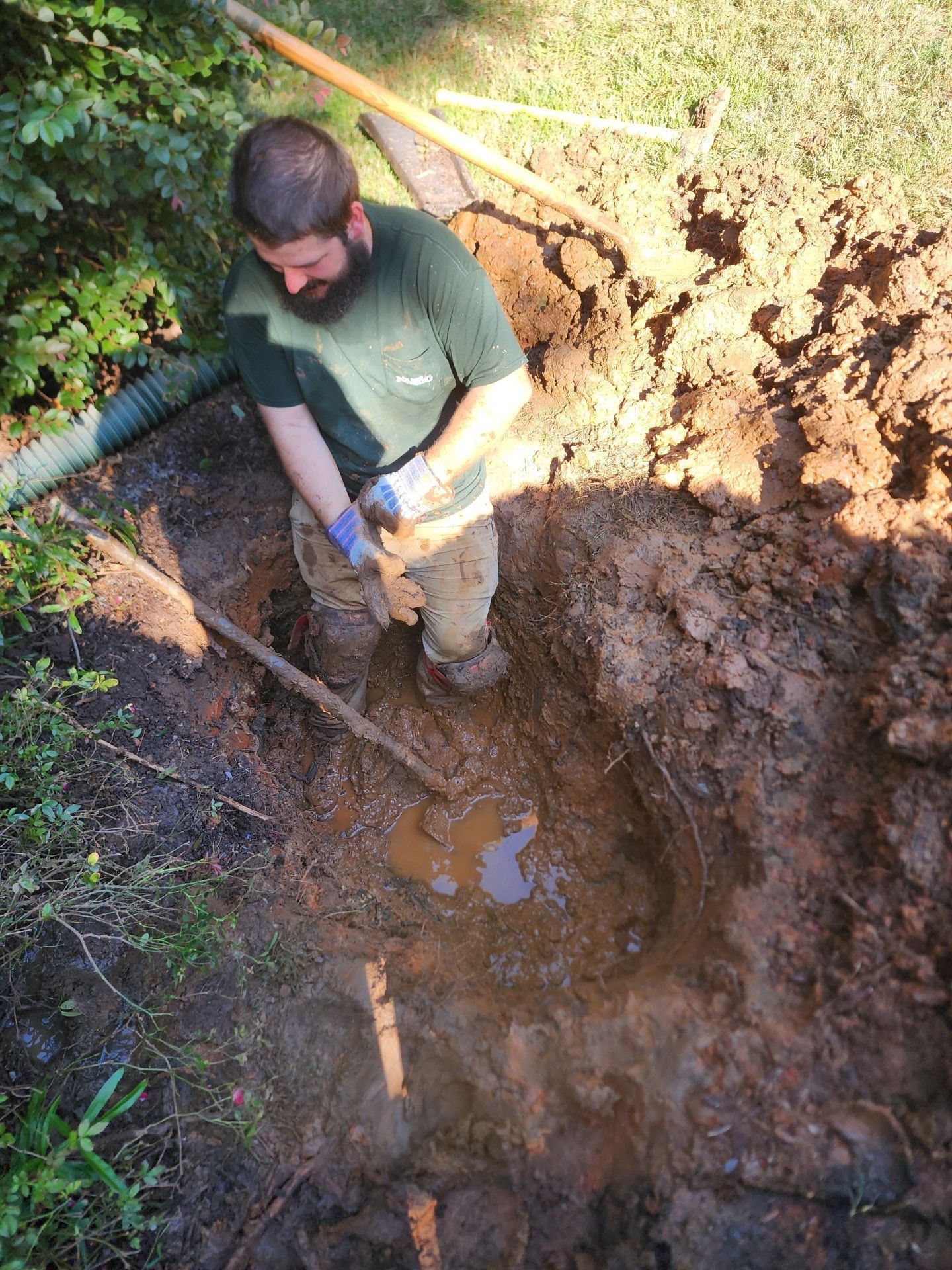 Man digging in a muddy hole outdoors. He wears gloves and a green shirt, using a shovel in the brown soil.