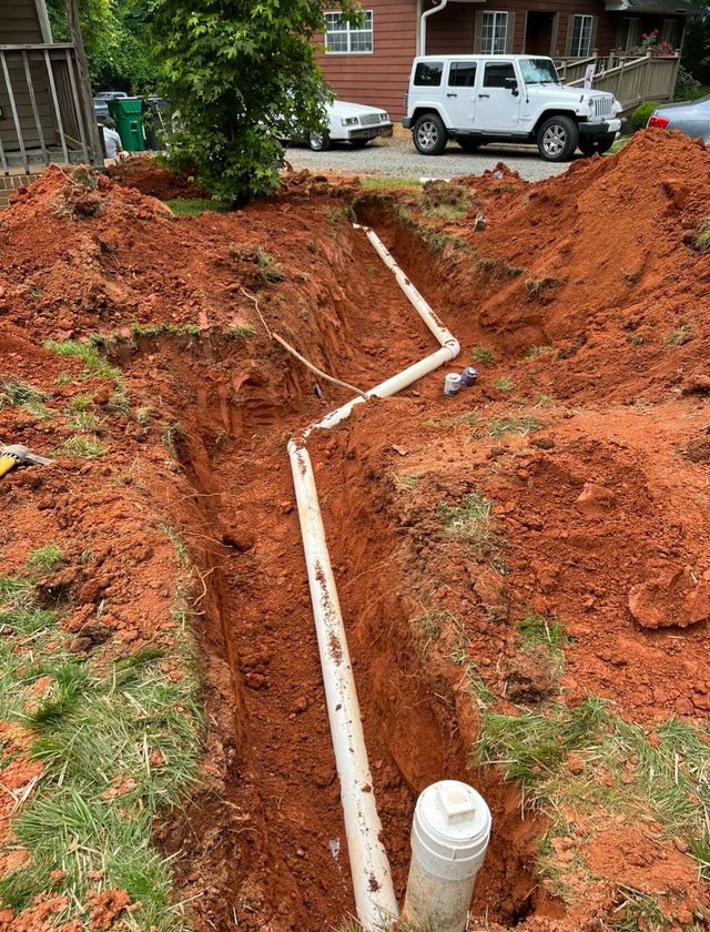 Trench dug in red soil, with white PVC pipes installed for plumbing, near a house and vehicles.