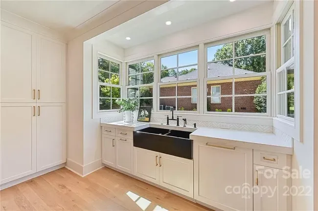 White kitchen with cabinetry, black sink, and large windows overlooking a residential neighborhood.