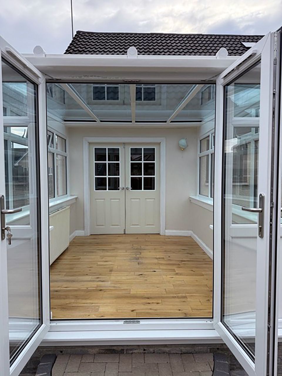 Conservatory with glass roof, white walls and doors, wooden floor and open French doors.