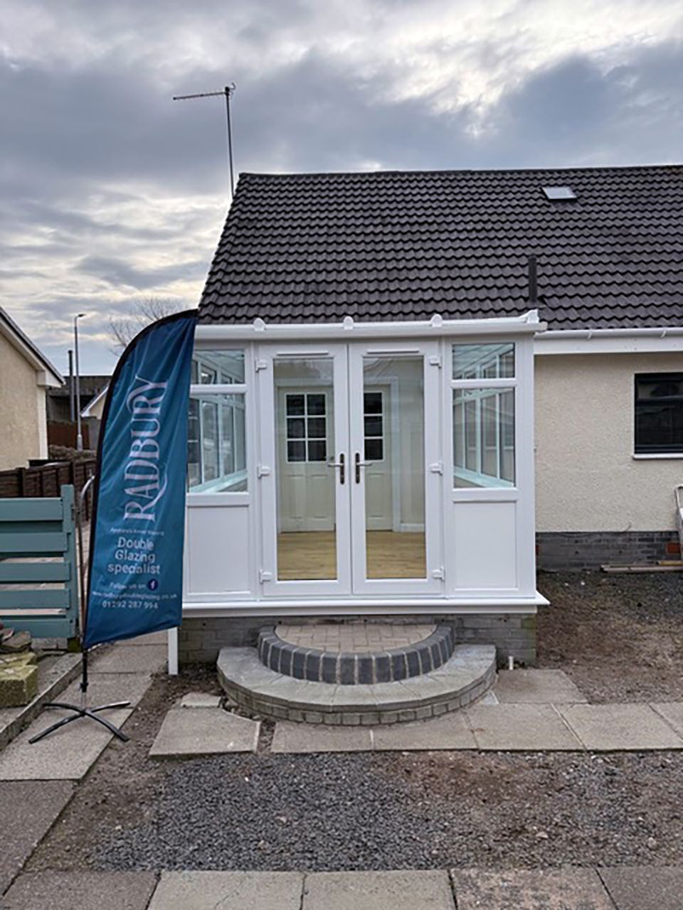 White conservatory with French doors, step, and banner in front of a house under a cloudy sky.