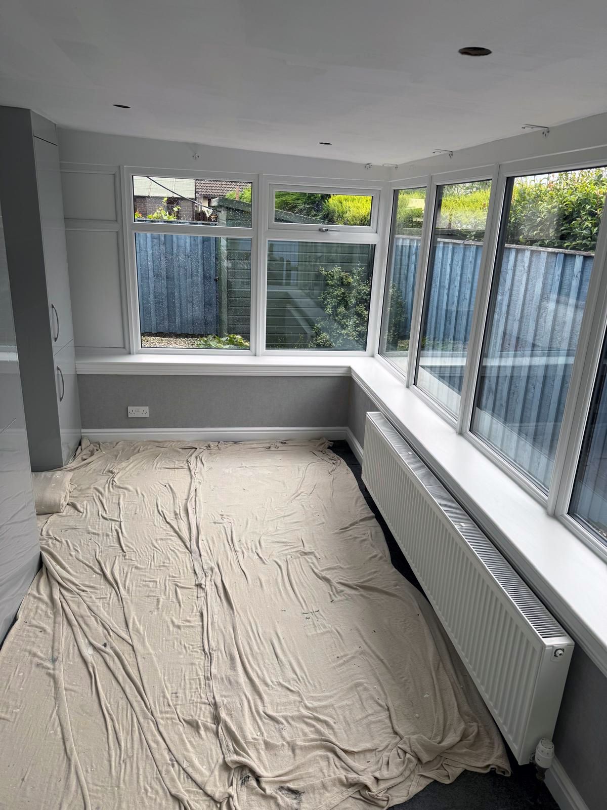 Empty sunroom with white trim, gray walls, and protective covering on the floor; windows offer outdoor views.