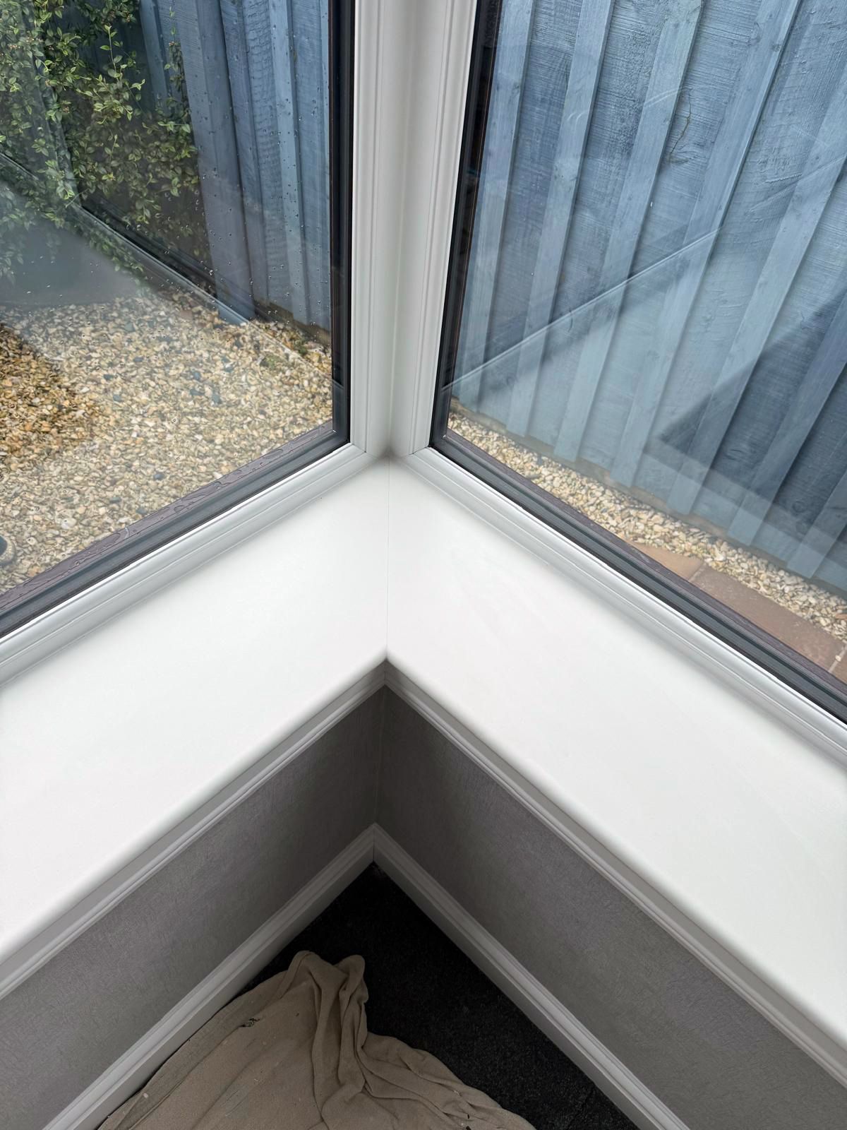 White window frame meeting at a corner, overlooking a gravel garden and a grey wooden fence.