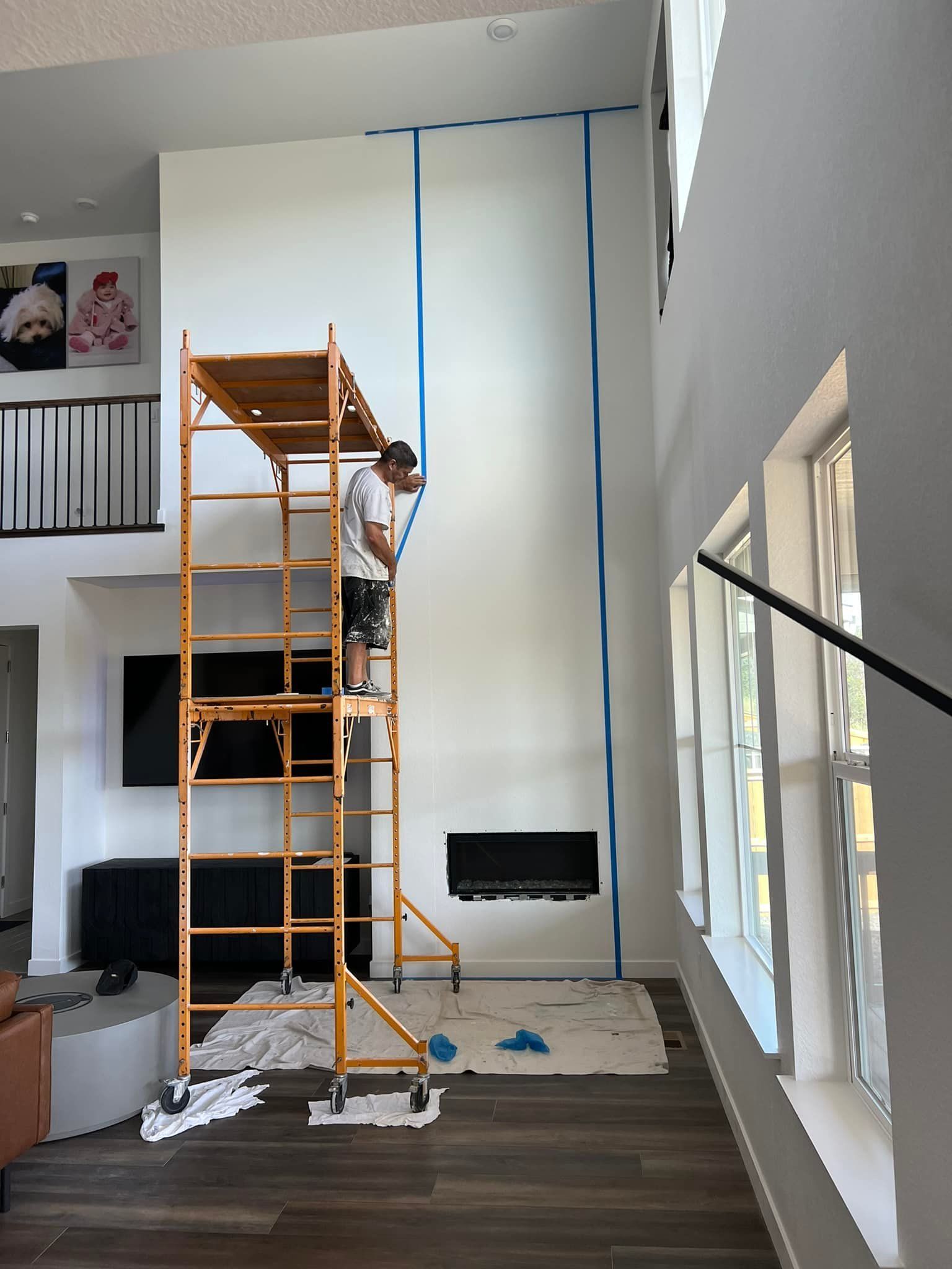 A man is standing on a scaffolding in a living room painting a wall.