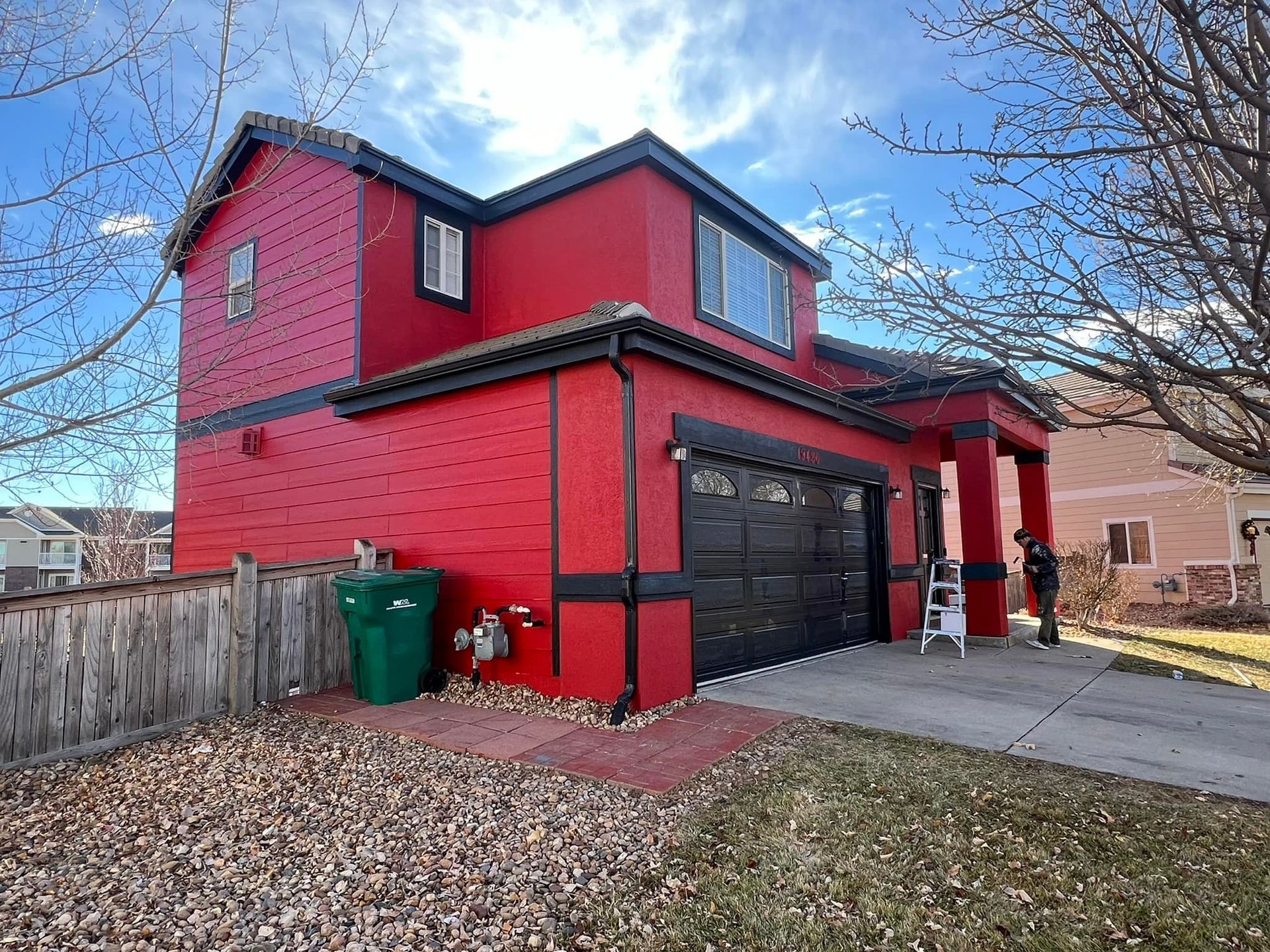 A red house with a black garage door and a green trash can in front of it.