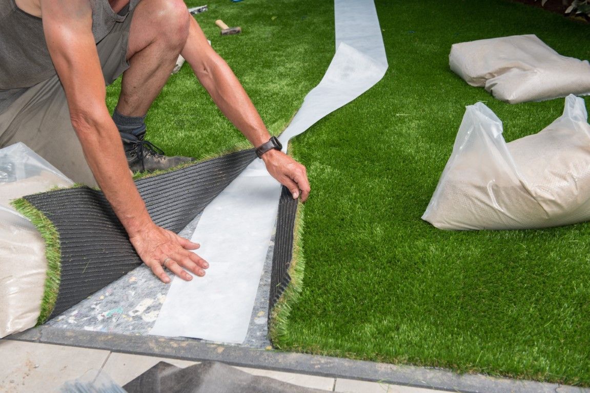 Man installs artificial turf, unrolling it over a white fabric, with bags of sand nearby.