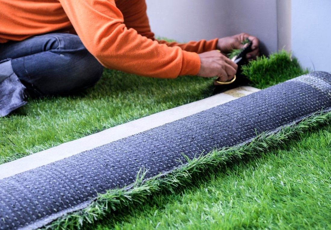 Person trimming artificial turf near a wall, using scissors. Turf roll visible.