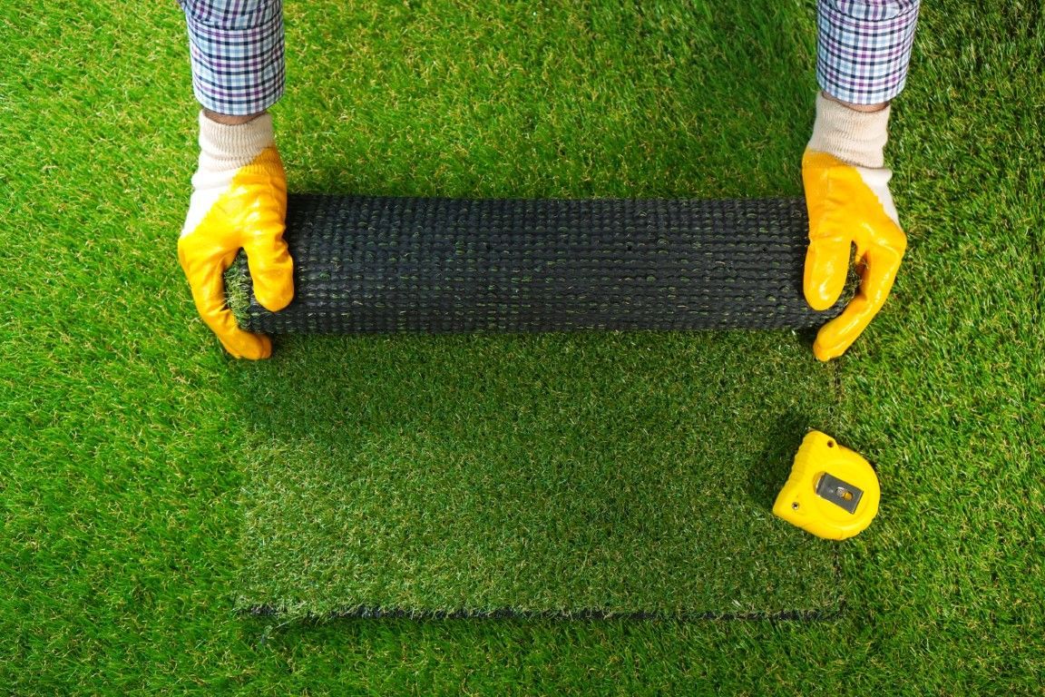 Person in yellow gloves unrolling black mesh on green artificial turf next to a yellow tape measure.