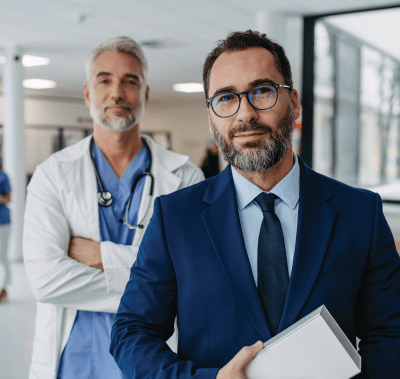 Man in suit stands in front of a doctor in a hallway, both looking at camera.