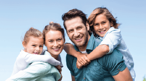 Family of four smiling outdoors, parents giving children piggyback rides under a blue sky.