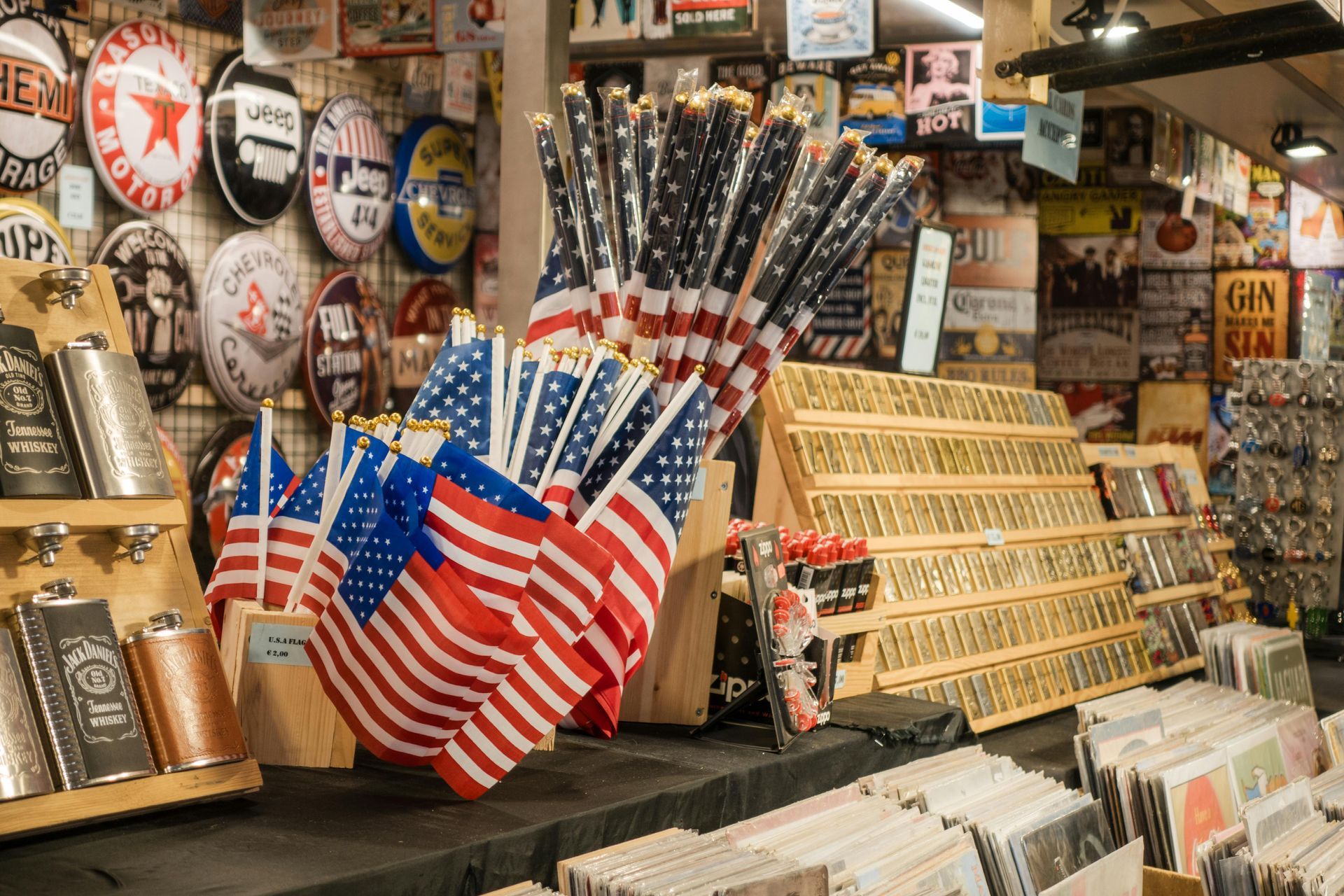 A cluster of small American flags sits on a counter in a shop filled with vintage-style signs, metal flasks, and records.