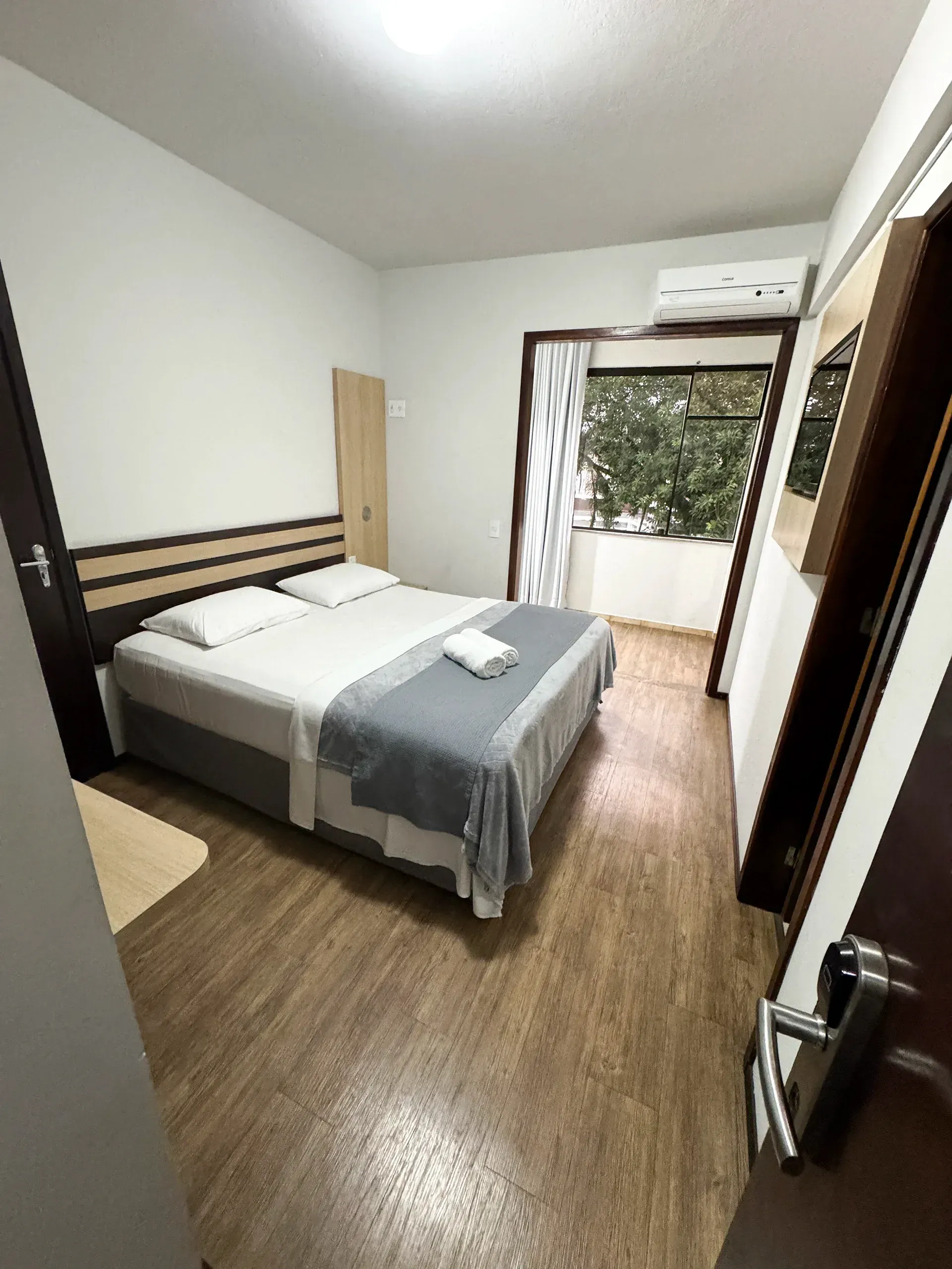 Quarto com cama de casal, piso de madeira, varanda com árvores, paredes brancas e ar condicionado.