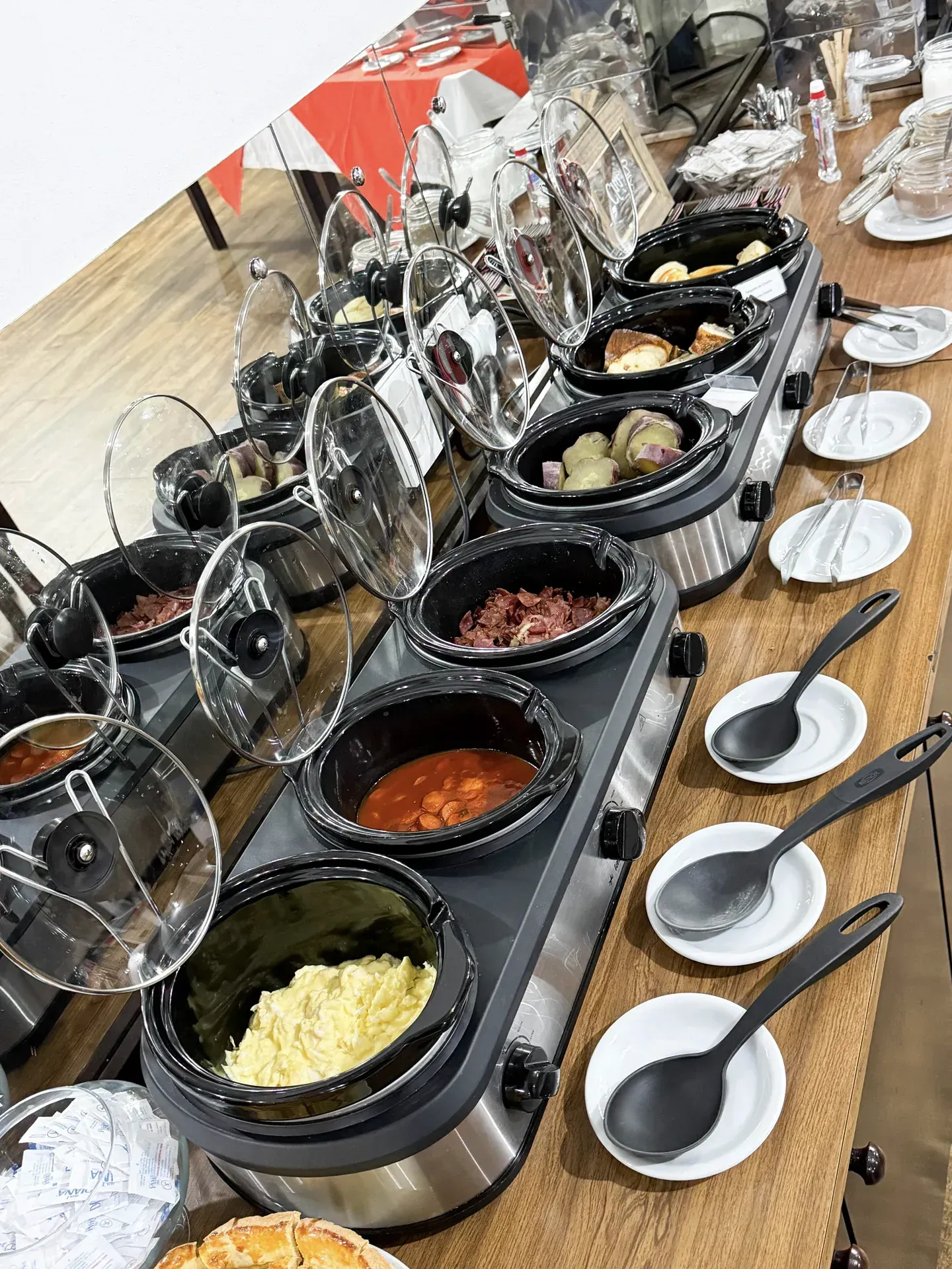 Mesa de buffet com várias panelas elétricas contendo alimentos, conchas e pratinhos.