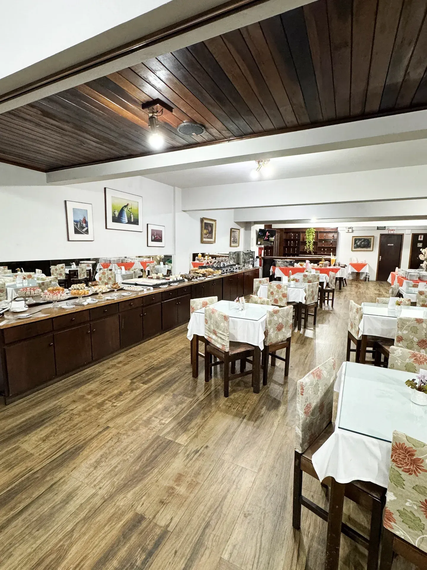 Área de jantar do restaurante com mesa de buffet, toalhas de mesa florais e teto de madeira.