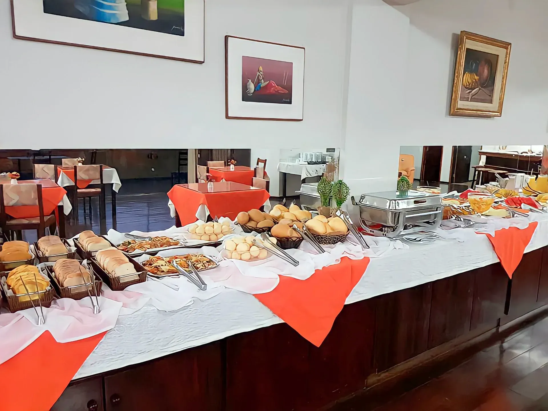 Buffet com diversos itens alimentícios em uma mesa comprida. Guardanapos vermelhos e renda branca sobre a mesa. Sala de jantar ao fundo.