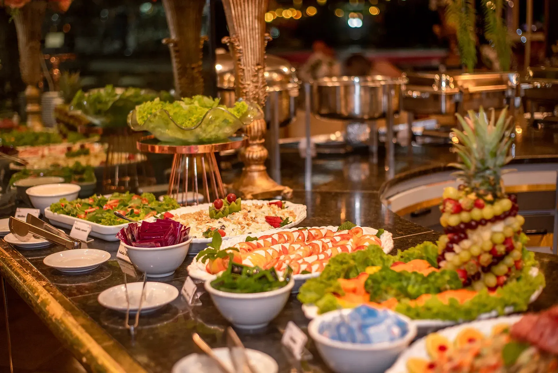 Mesa de bufê com diversas saladas e guarnições, incluindo um abacaxi decorado com frutas.