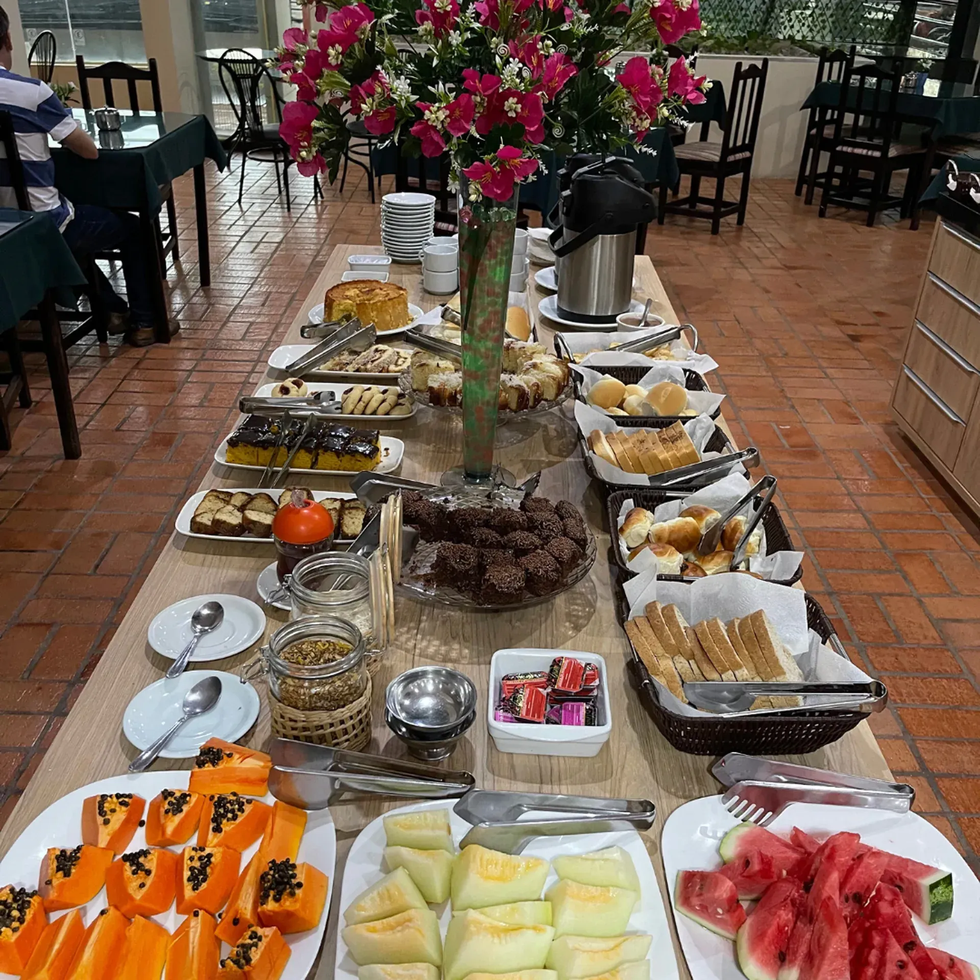 Mesa de bufê com diversos pães, doces e uma torta em um ambiente de restaurante.