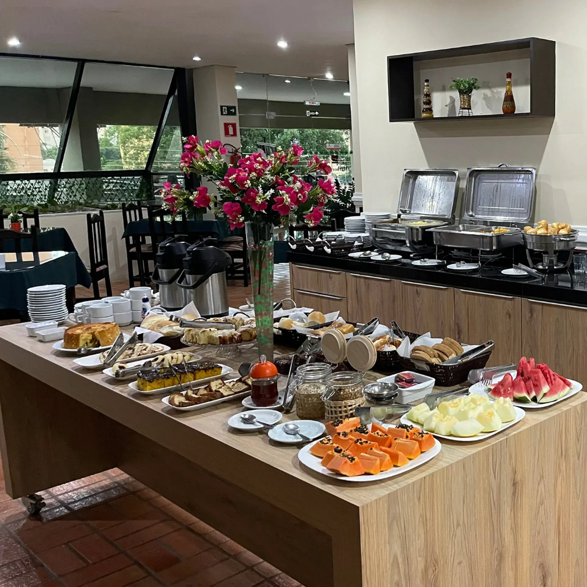 Área de jantar do restaurante com mesa de buffet, toalhas de mesa florais e teto de madeira.