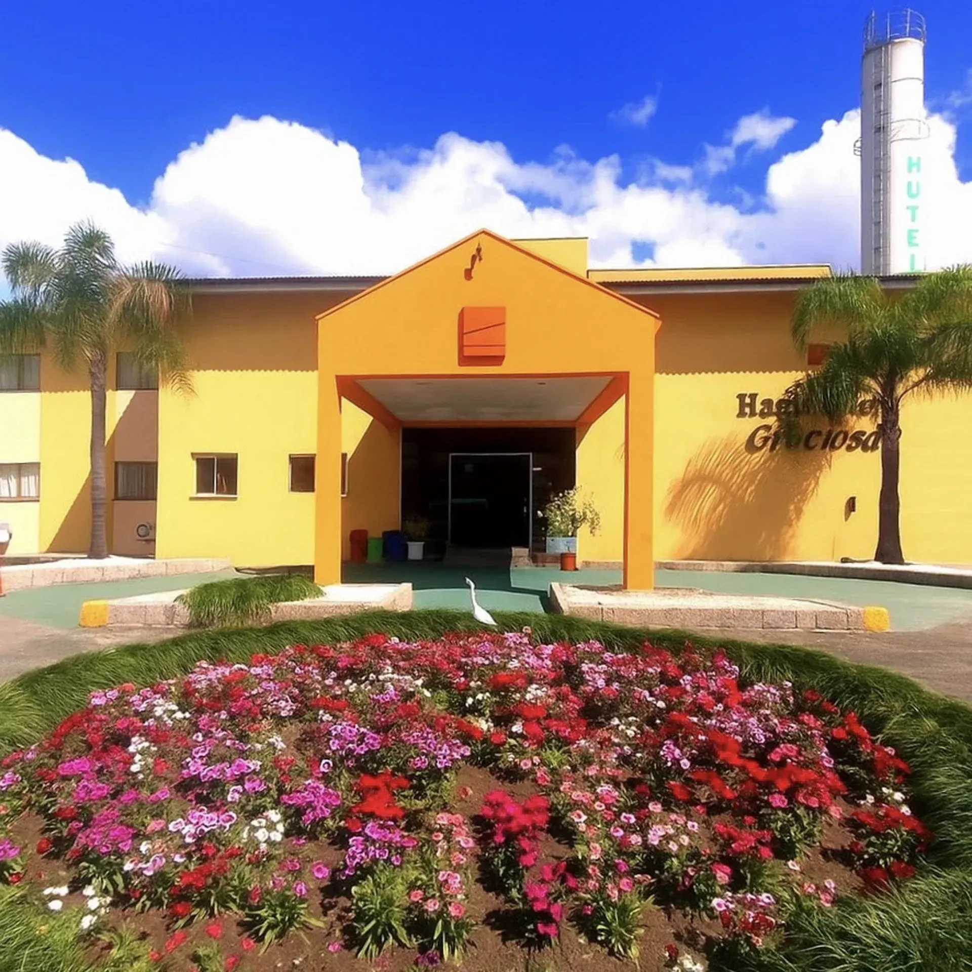 Prédio azul e branco com um amplo estacionamento, palmeiras e uma rua em frente. Dia ensolarado.