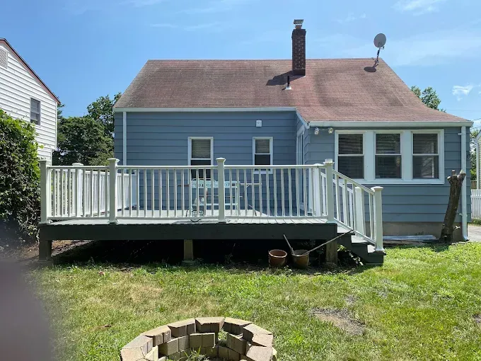 Blue house with a white deck, brown roof, and brick fire pit in a grassy yard under a blue sky.