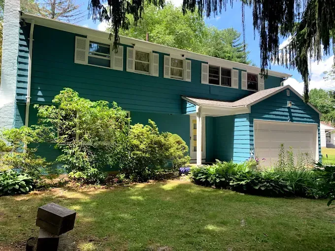 Two-story teal house with white trim, shutters, and a garage, surrounded by greenery.