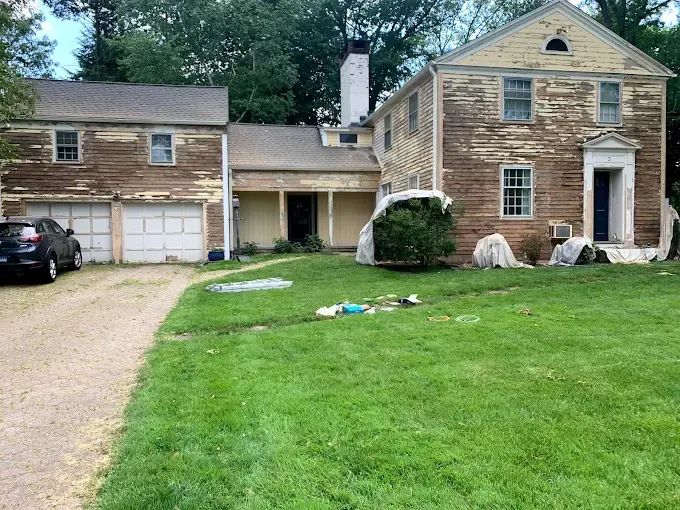 House with peeling paint and three-car garage, with overgrown lawn and gravel driveway.