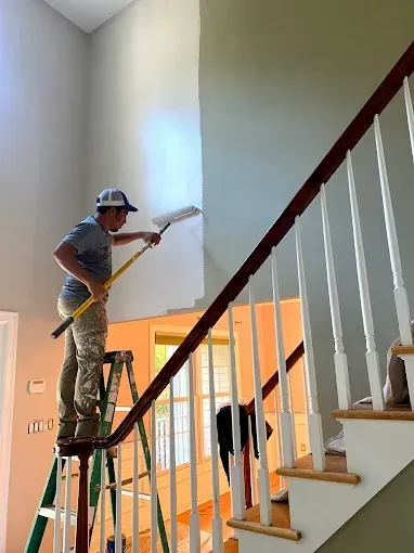 Person painting a high wall above a staircase. They are using a roller while standing on a ladder.
