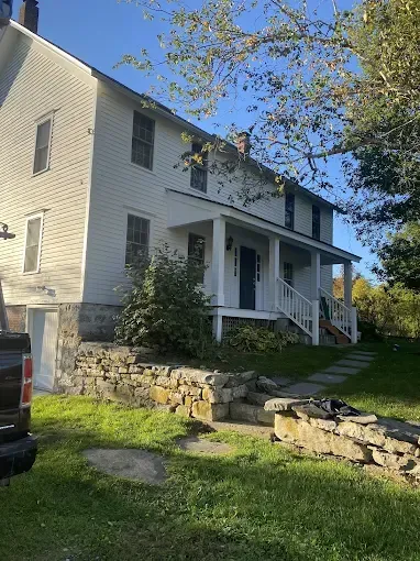 White, two-story house with a porch and stone foundation. Lawn in front with stone retaining walls. Sunlight.