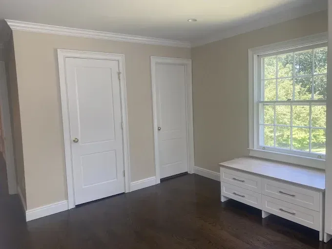 Empty room with two white doors, a window with a bench, and dark wood floors.