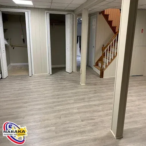 Finished basement with gray wood-look flooring, beige walls, support beams, stairs, and open doorways.