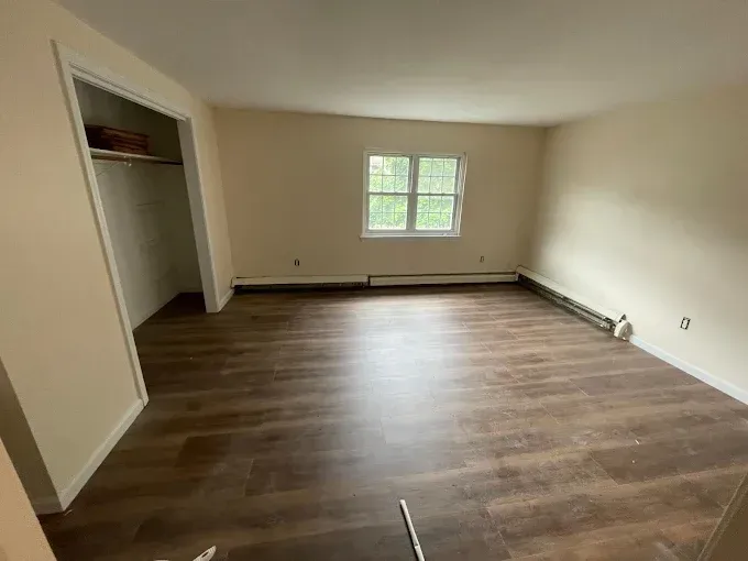 Empty room with brown flooring, beige walls, small window, and open closet.