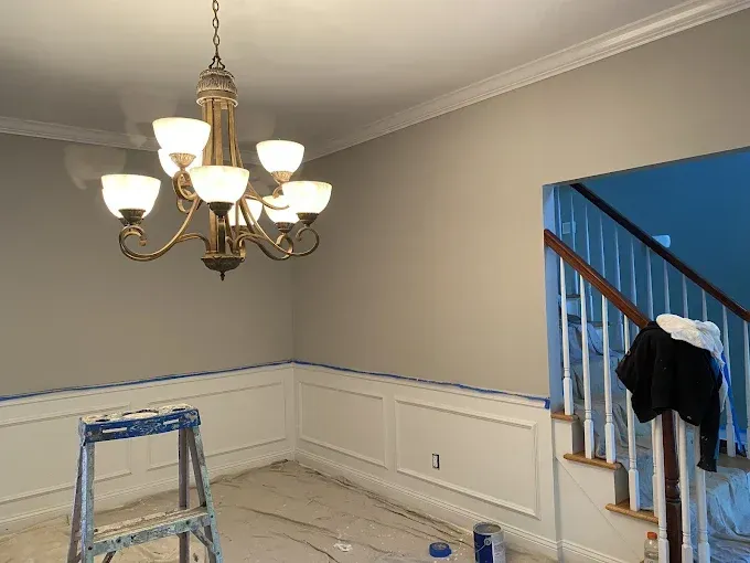 Room with gray walls, white wainscoting, and a brass chandelier; a staircase is in the adjacent room.